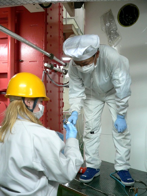 Ochi-san, standing on a short platform, bends at the waist to receive the 3 cm cylindrical plug from Juli, looking stunning in her yellow hard hat. (84K JPEG)