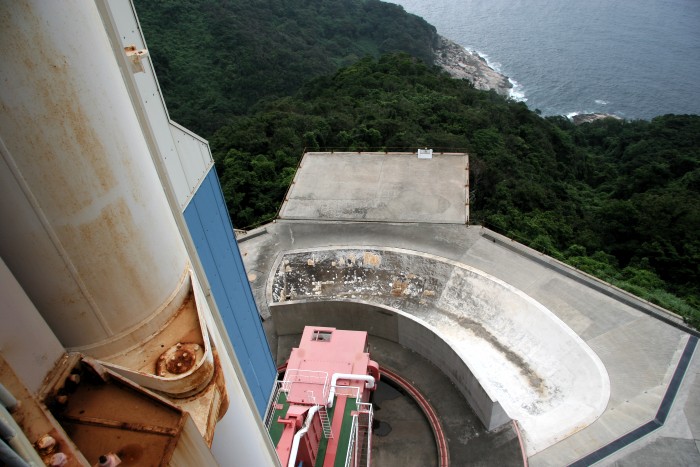 The massive bulk of a giant hinge dominates the left side of the photo, as the lines of the door fall away to the ground, scarily distant.  Beyond the gray concrete of the flame deflector and pad area the dark green foliage tumbles down to the deep blue sea. (94K JPEG)