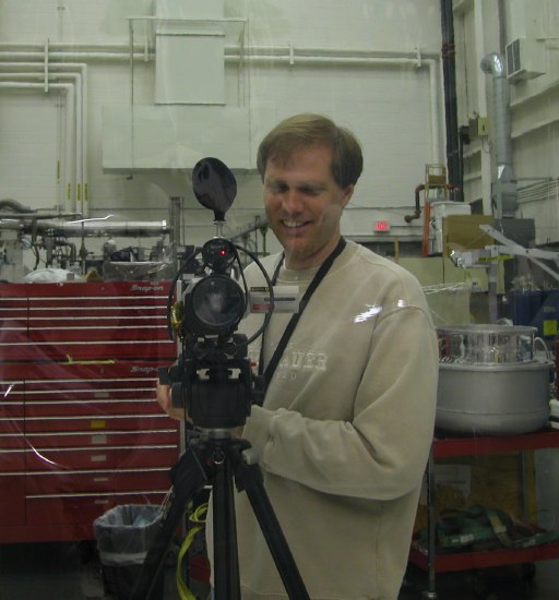The shotgun mic, tally lamp, and 57mm lens of a video camera stare directly at us like the eye of some three-legged monster, as the camera operator grins evilly. (52K JPEG)